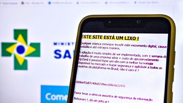 Um smartphone mostra uma mensagem deixada por um hacker em ataque ao site do Ministério da Saúde, mostrado no fundo. - Sputnik Brasil
