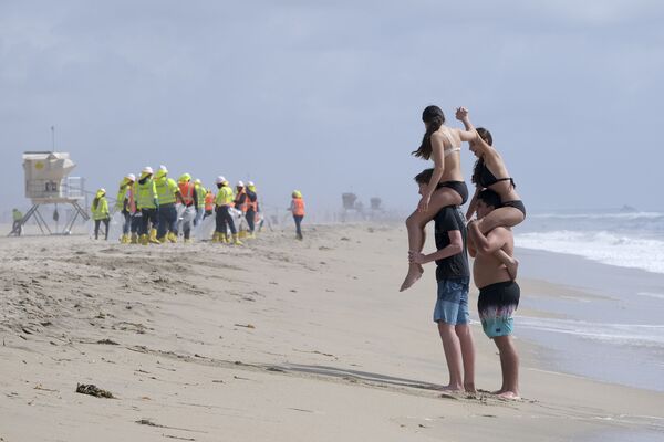 Pessoas se divertem enquanto trabalhadores em trajes de proteção limpam a praia poluída em Huntington Beach, Califórnia, EUA. - Sputnik Brasil