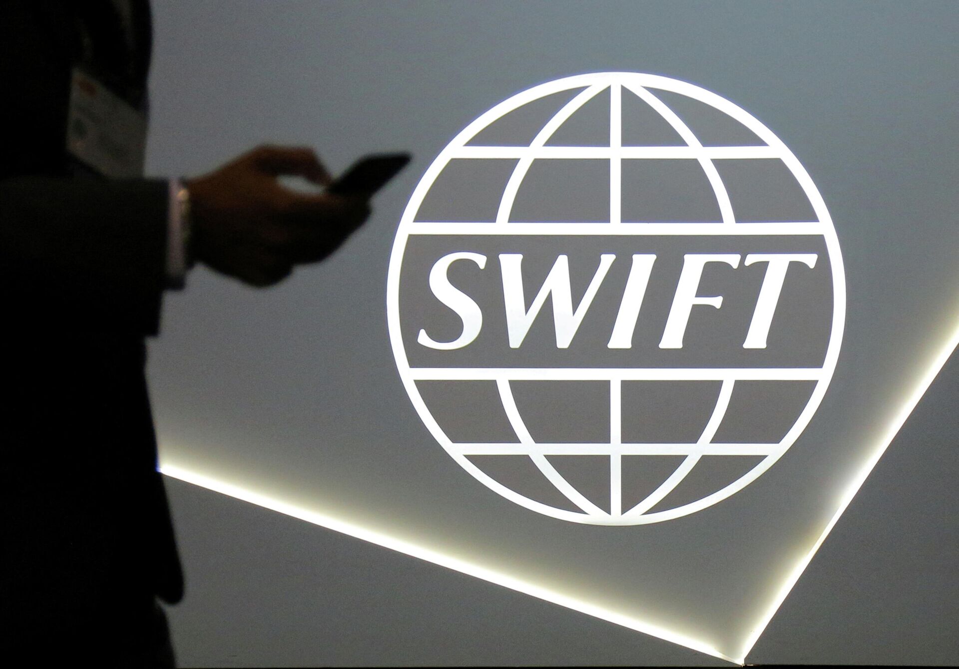 Um homem usando um telefone celular passa pelo logotipo da Sociedade Mundial de Telecomunicações Financeiras Interbancárias (SWIFT, na sigla em inglês) na conferência bancária e financeira SIBOS em Toronto, Ontário, Canadá, 19 de outubro de 2017 - Sputnik Brasil, 1920, 15.03.2022