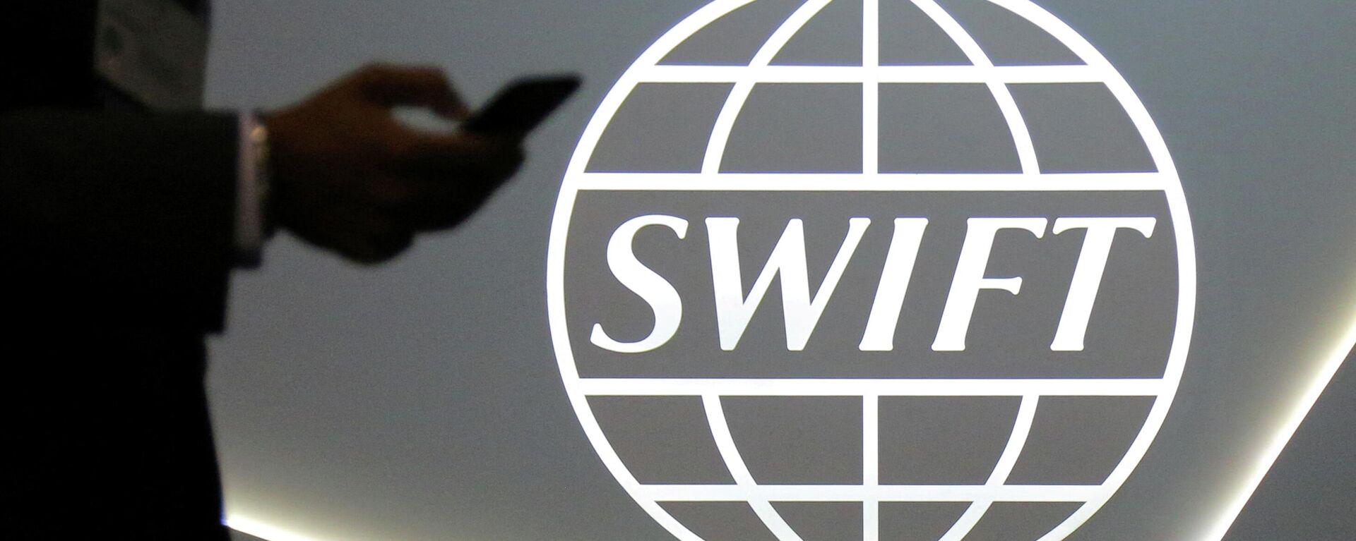 Um homem usando um telefone celular passa pelo logotipo da Sociedade Mundial de Telecomunicações Financeiras Interbancárias (SWIFT, na sigla em inglês) na conferência bancária e financeira SIBOS em Toronto, Ontário, Canadá, 19 de outubro de 2017 - Sputnik Brasil, 1920, 28.02.2022