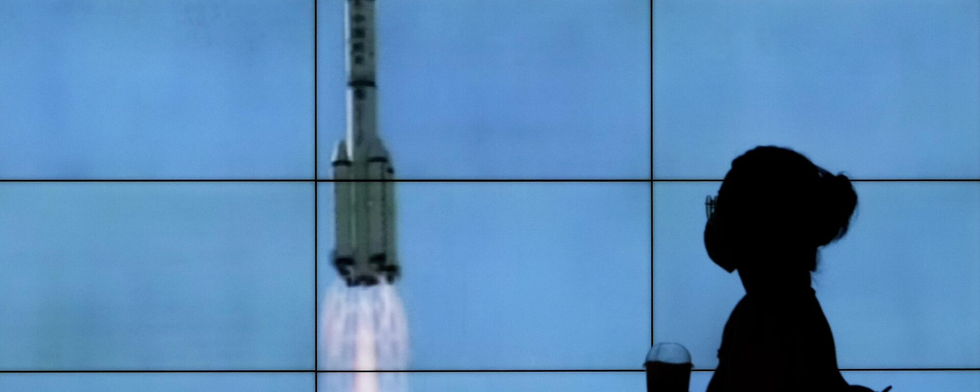 Uma pessoa de máscara caminha em frente a uma tela mostrando um lançamento do foguete Longa Marcha-2F Y12 em uma transmissão da CCTV, em Pequim, China, 17 de junho de 2021 - Sputnik Brasil, 1920, 05.06.2022