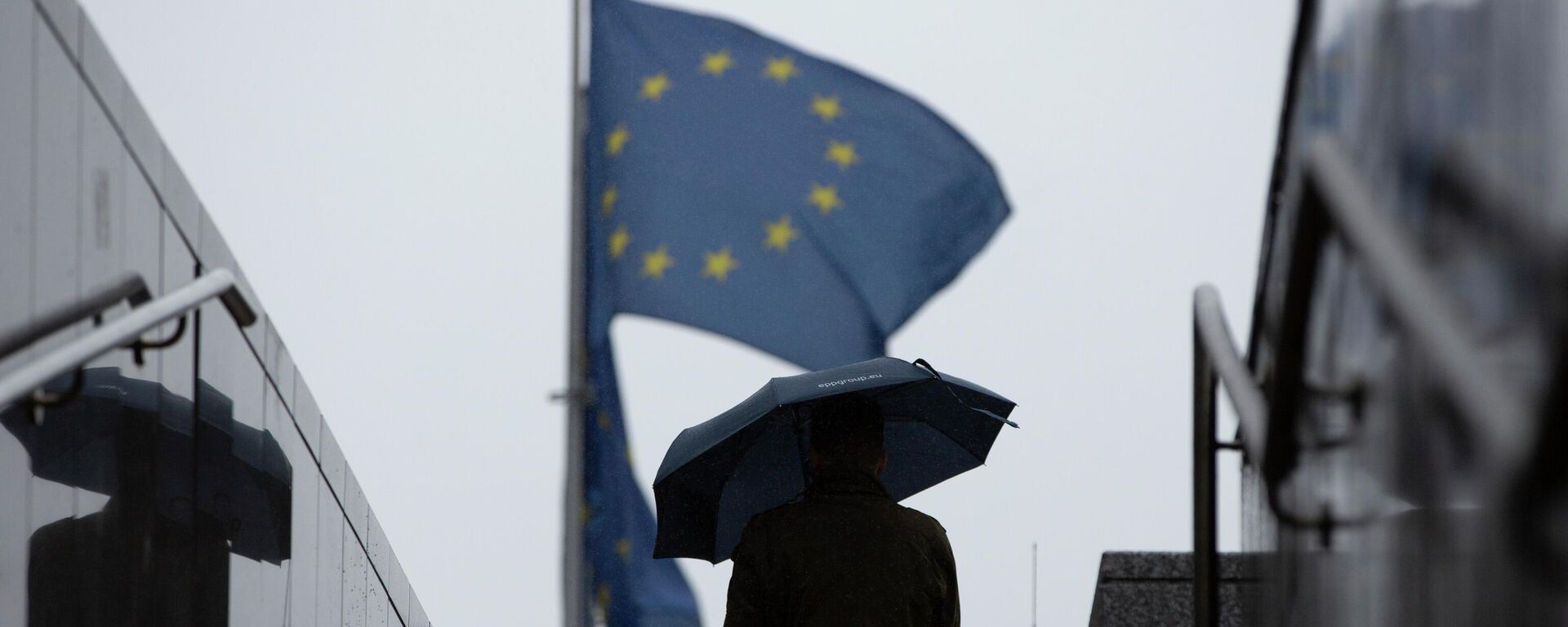 Homem caminha com guarda-chuva perto de bandeiras da União Europeia no exterior da sede do bloco, em Bruxelas, na Bélgica, em 16 de outubro de 2019 (foto de arquivo) - Sputnik Brasil, 1920, 13.09.2022