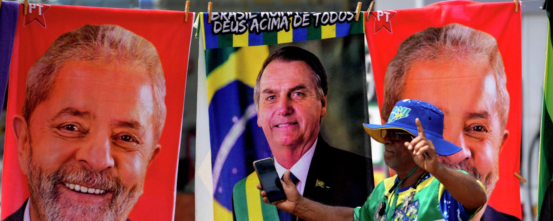 Apoiador vestido com as cores da bandeira do Brasil passa em frente a toalhas à venda com os rostos dos candidatos à Presidência Jair Bolsonaro (PL) e Luiz Inácio Lula da Silva (PT). Brasília, 23 de setembro de 2022 - Sputnik Brasil, 1920, 13.10.2022