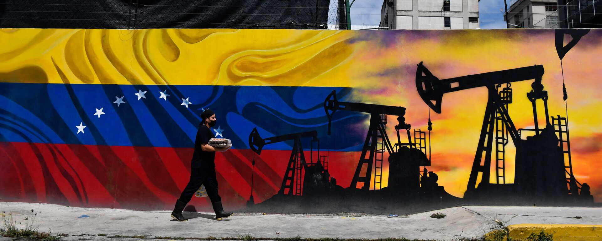 Um homem usando uma máscara facial passa por um mural representando uma bomba de petróleo e a bandeira venezuelana, em uma rua de Caracas, em 26 de maio de 2022 - Sputnik Brasil, 1920, 30.05.2023