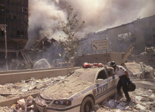 Um resgatador entra em um carro da Polícia de Nova York coberto de escombros enquanto bombeiros da cidade pulverizam água em ruínas fumegantes em fundo, após o ataque terrorista de 11 de setembro no World Trade Center. - Sputnik Brasil