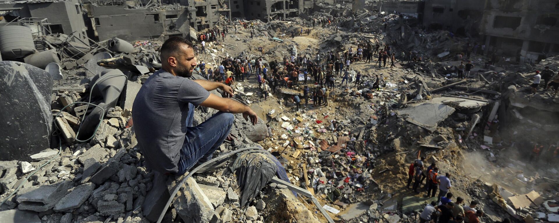 Homem senta-se sobre os escombros enquanto outros passeiam entre os destroços de edifícios alvos de ataques aéreos israelenses no campo de refugiados de Jabaliya, no norte da Faixa de Gaza, 1º de novembro de 2023 - Sputnik Brasil, 1920, 09.12.2023