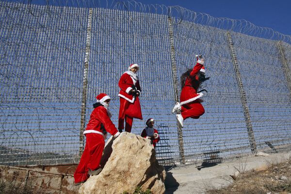 Palestinos vestidos de Papai Noel pulando de uma pedra ao lado de uma cerca com arame farpado de uma barreira de segurança feita por Israel perto do vilarejo de Wallajeh - Sputnik Brasil