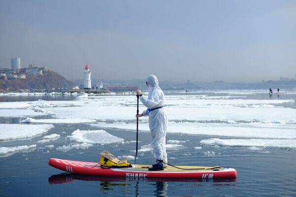 Homem com traje de proteção contra coronavírus sobre prancha em águas de Vladivostok, Rússia, 21 de março de 2020 - Sputnik Brasil