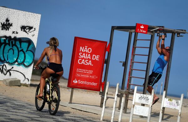 Mulher passa de bicicleta perto de um cartaz que diz Não saia de casa na praia de Ipanema, no Rio de Janeiro, durante a epidemia de coronavírus - Sputnik Brasil
