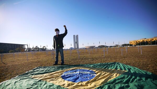 Ativista em frente à bandeira brasileira com cruzes no fundo que simbolizam vítimas da COVID-19 durante protesto contra Bolsonaro em Brasília, 28 de junho de 2020. - Sputnik Brasil